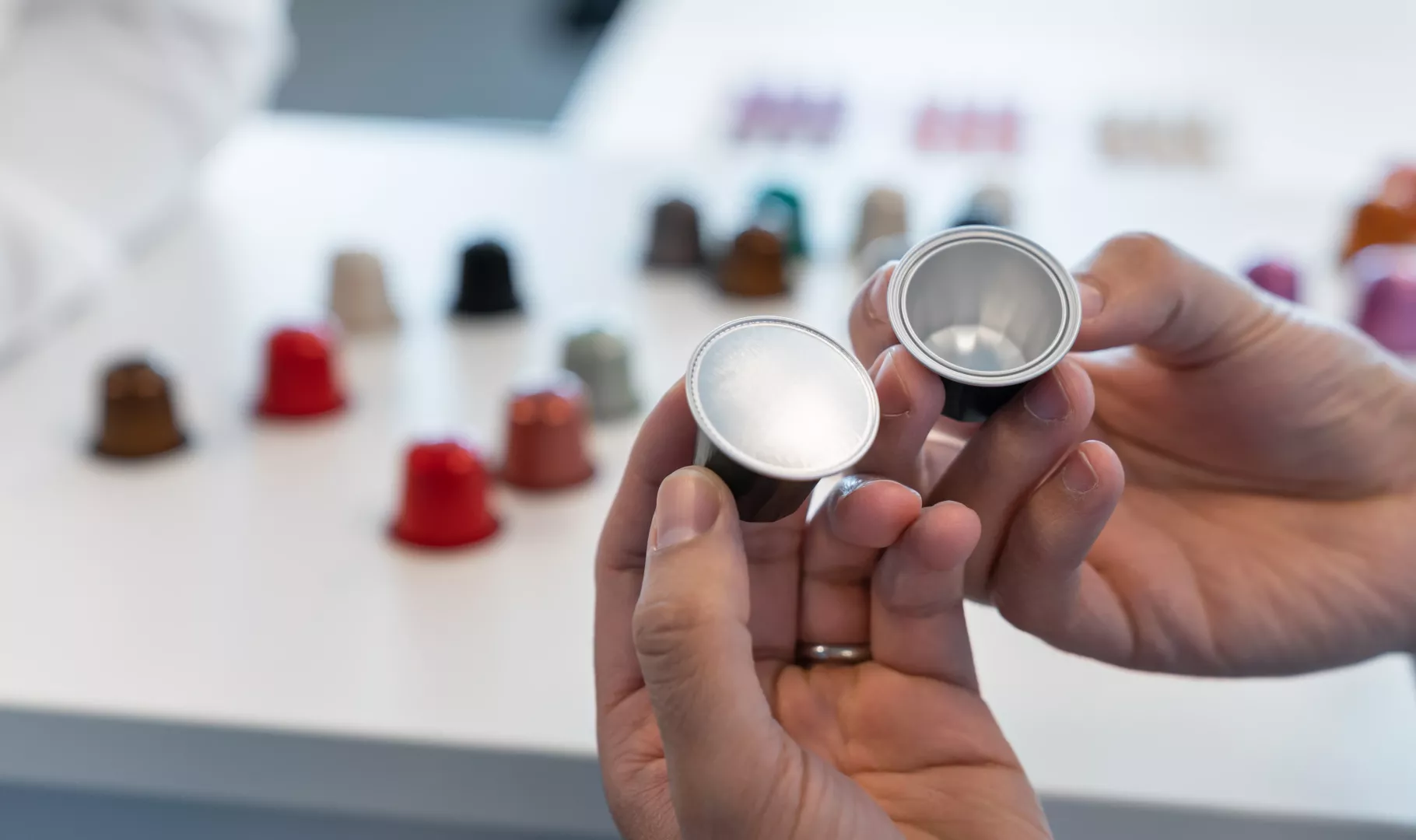 Hands holding two empty AluSense aluminium coffee capsules, with a range of coloured capsules in the background.