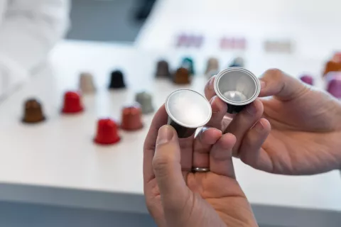 Hands holding two empty AluSense aluminium coffee capsules, with a range of coloured capsules in the background.